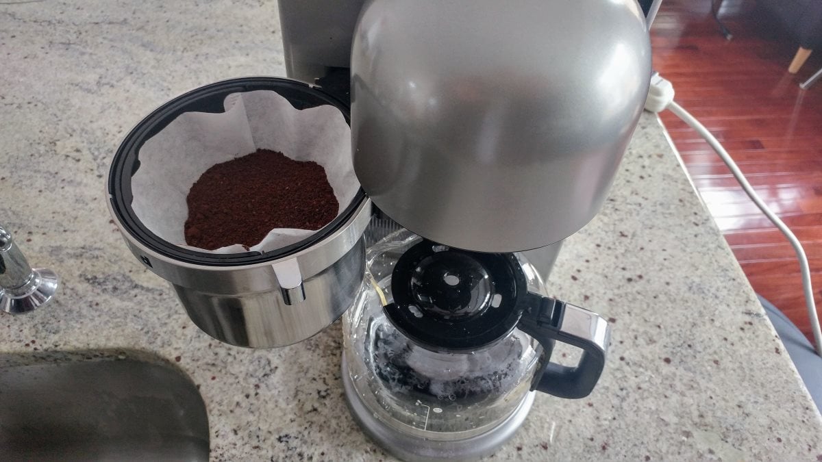 A top-down view of the KitchenAid KCM0802 coffeemaker. The brew basket is open, revealing that it is lined with a paper filter and about half full of ground coffee.