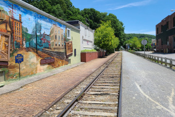 A railroad is bordered by a brick walkway on the left and a wide gravel bike path on the right. A mural next to the trail depicts the history of Glen Rock.
