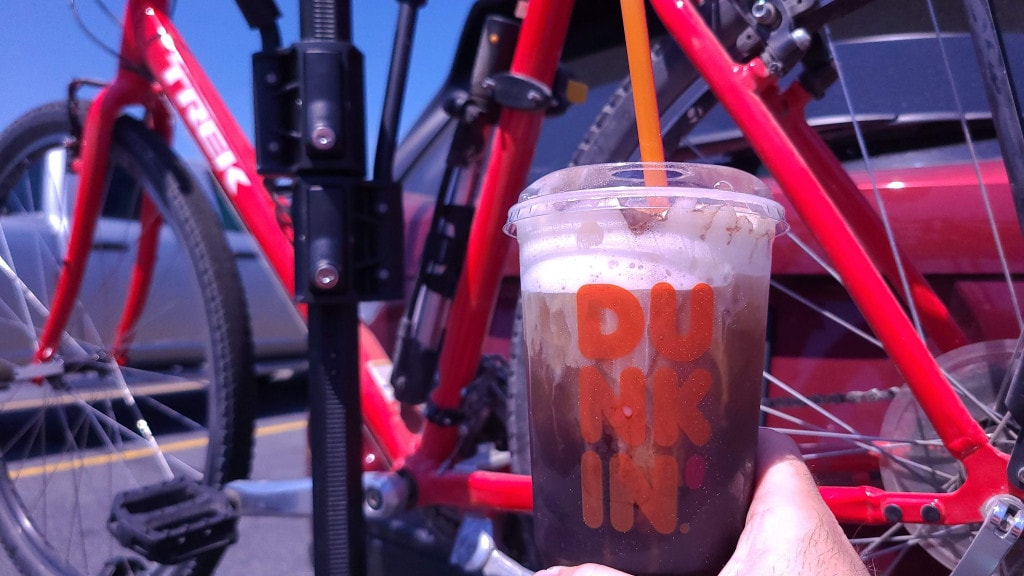 A hand holds a cup of iced coffee in front of a red Trek bike hanging on the back of a red SUV. The cup says "Dunkin".